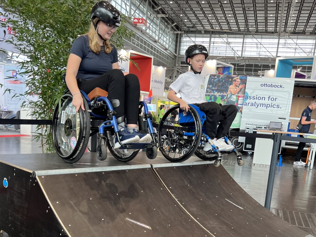 Eine Rollstuhlfahrerin und ein Junge im Rollstuhl stehen gemeinsam auf den Hinterrädern balancierend auf einer Quarter Ramp in einer Messehalle. 