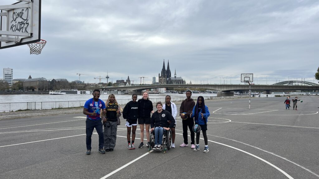 Eine kleine Gruppe von Menschen stehen gemeinsam mit David, der der einzige Rollstuhlfahrer ist auf einem Basketballfeld direkt am Rhein. Im Hintergrund sieht man den Kölner Dom. Die Gruppe besteht aus Menschen aus Tansania und Deutschland.