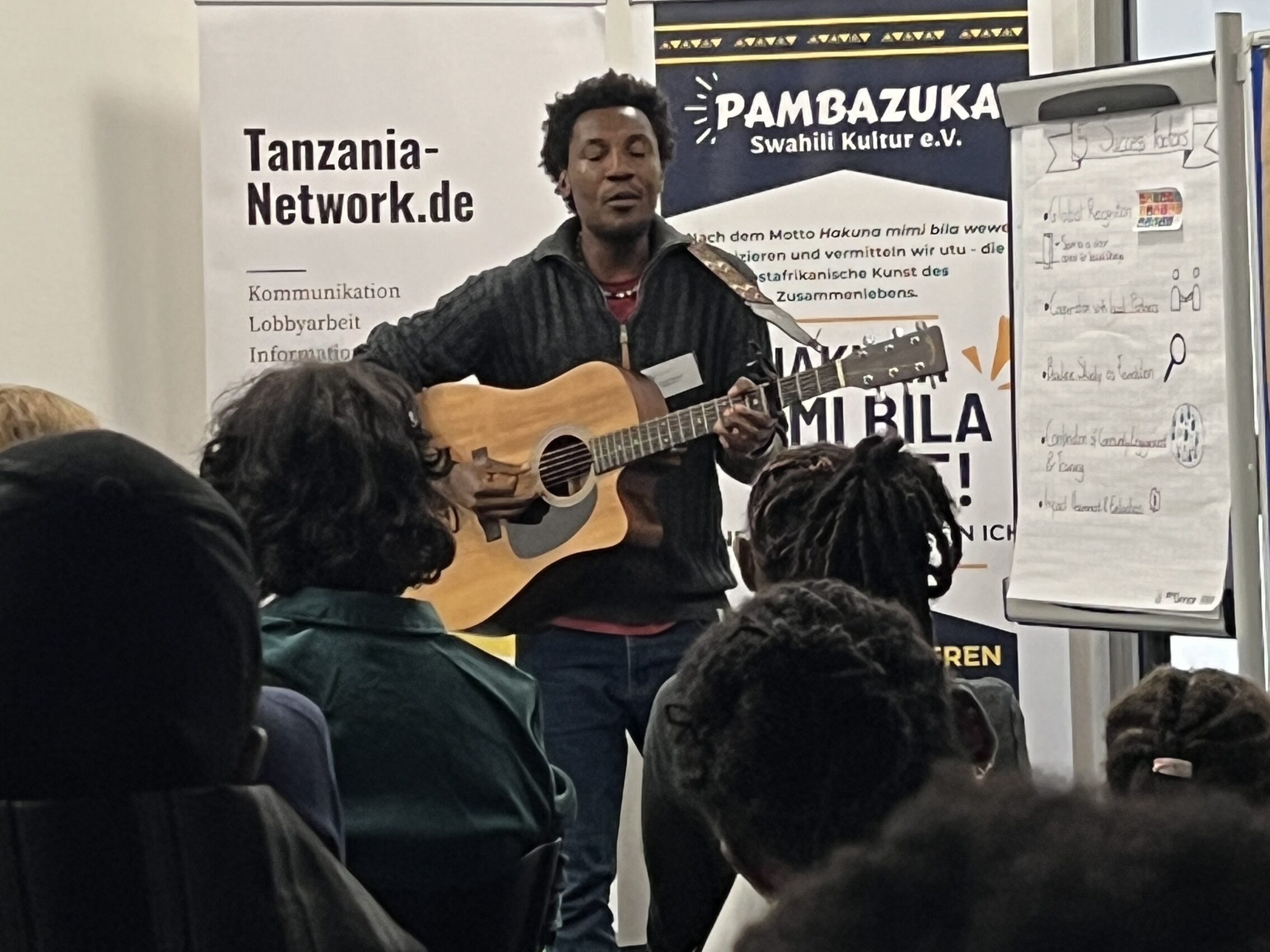 Ein transsporadischer Sänger steht mit einer Akustikgitarre vor den Zuschauern. Im Hintergrund Banner von den Organisatoren des Tansania Netzwerk und Pambazuka e.V.