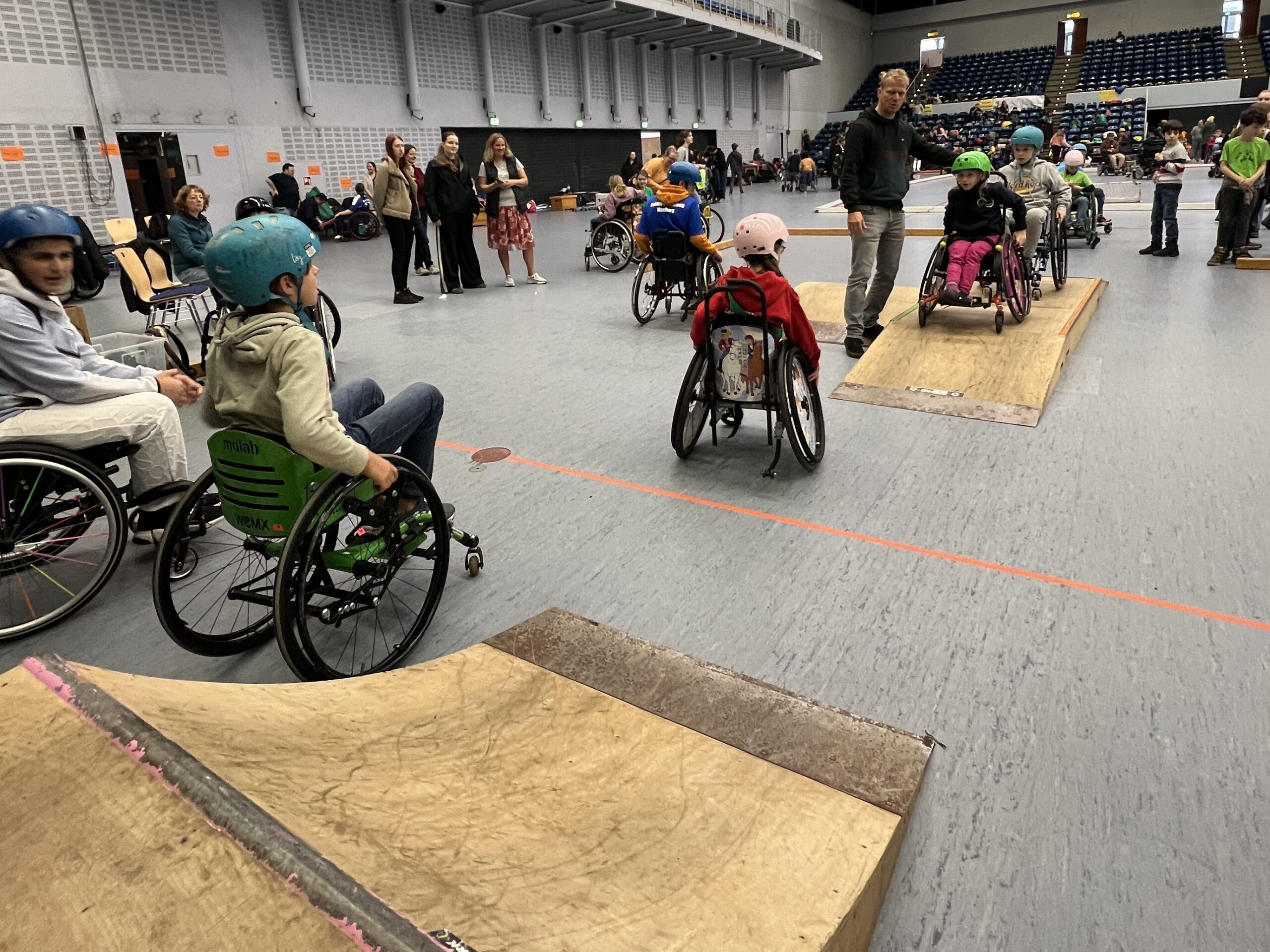 in einer Sporthalle sind viele Kinder im Rollstuhl und fahren über einen Skatepark aus mobilen Holzelementen.