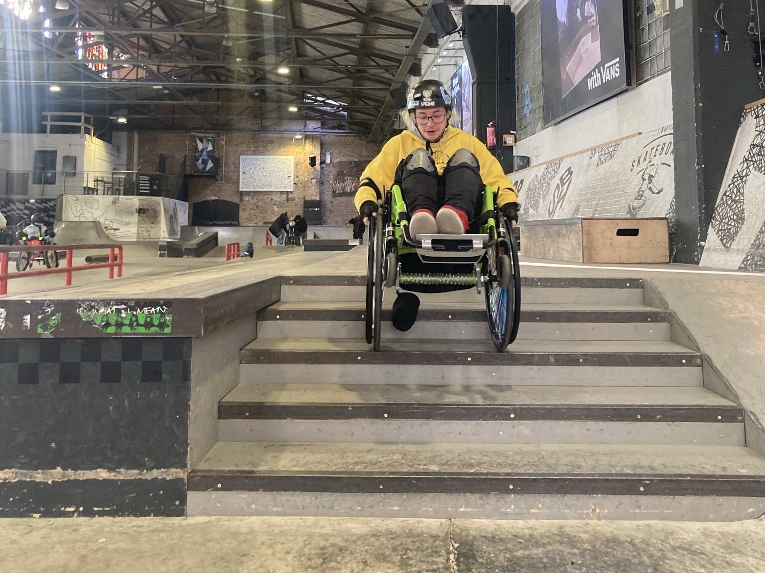 Eine Person im Rollstuhl fährt eine Treppe in einer Skatehalle hinunter. Sie trägt einen gelben Hoodie und einen Helm, der das Gesicht schützt. Die Skatehalle ist groß, mit hohen Decken und Metallstreben, die die Konstruktion stützen. Im Hintergrund sind Rampen, Rails und weitere Skater*innen zu sehen, die den Raum nutzen. Die Szene zeigt die aktive Nutzung der Halle und die Herausforderungen des WCMX-Sports.
