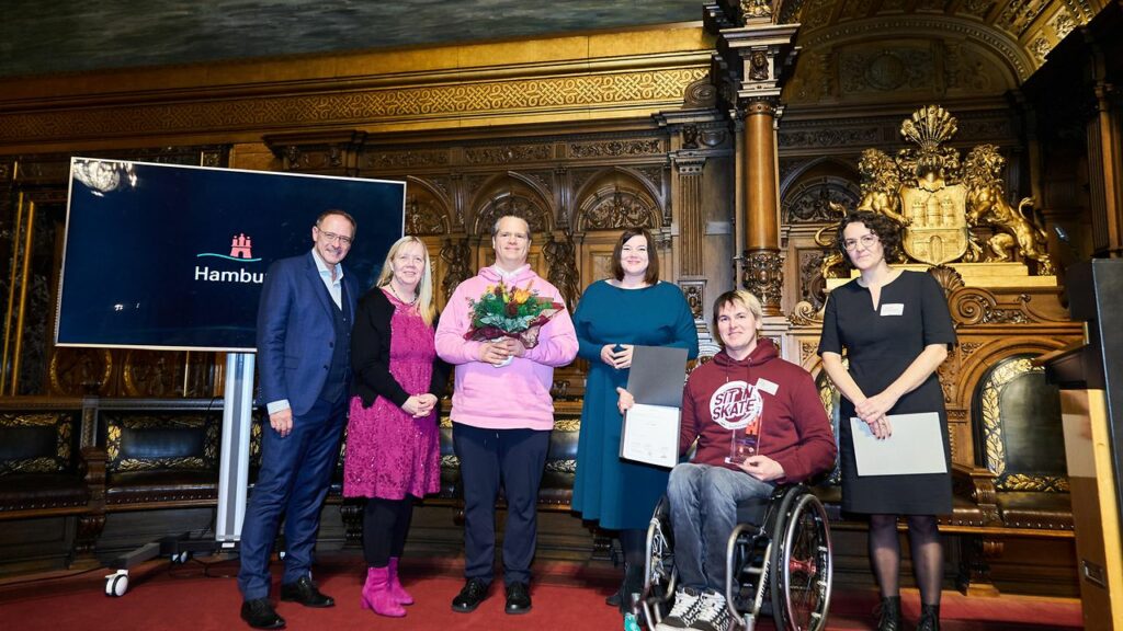 Auf einer Bühne stehen drei Frauen und drei Männer, unter ihnen Graham und David von SIT'N'SKATE. David ist Rollstuhlfahrer. Sie sehen festlich aus und halten Blumenstrauß, Auszeichnung und Urkunde in der Hand.