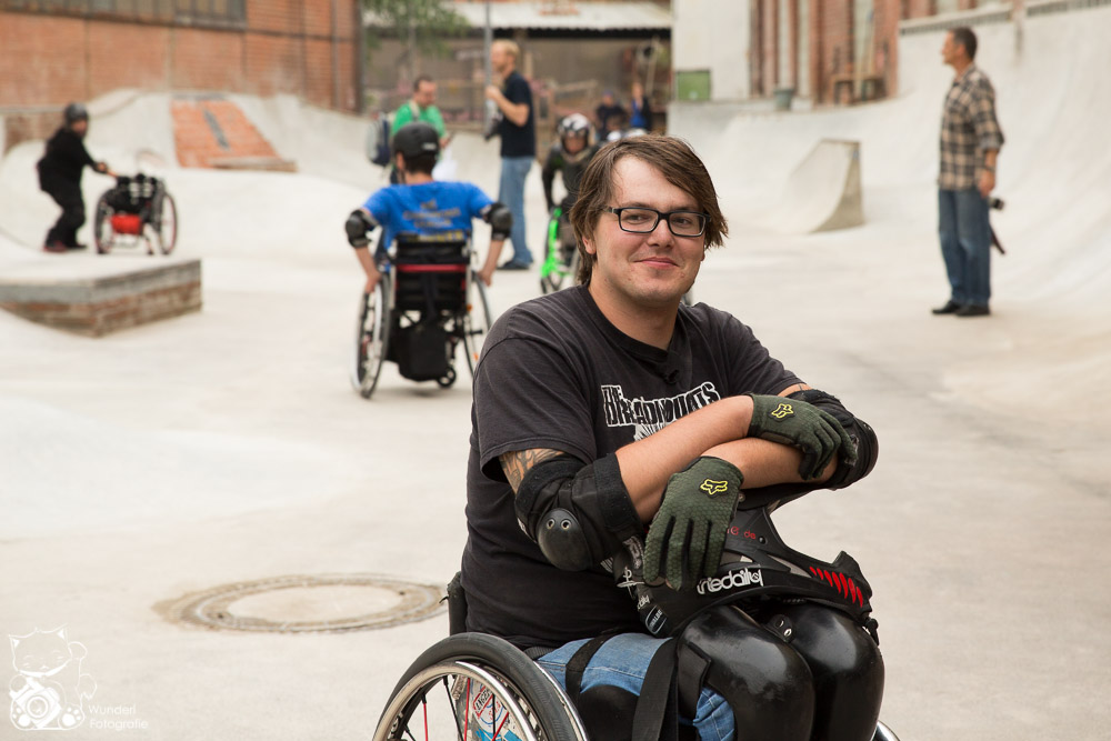 David Lebuser steht mit seinem Rollstuhl im Skatepark an der Skatehalle Mr Wilson in Kassel. Seinen Helm auf den Schoß, er hat mittellange Haare, eine Brille und seine Arme über kreuz auf dem Helm. Im Hintergrund Betonrampen und weitere Rollstuhlfahrende im Skatepark.